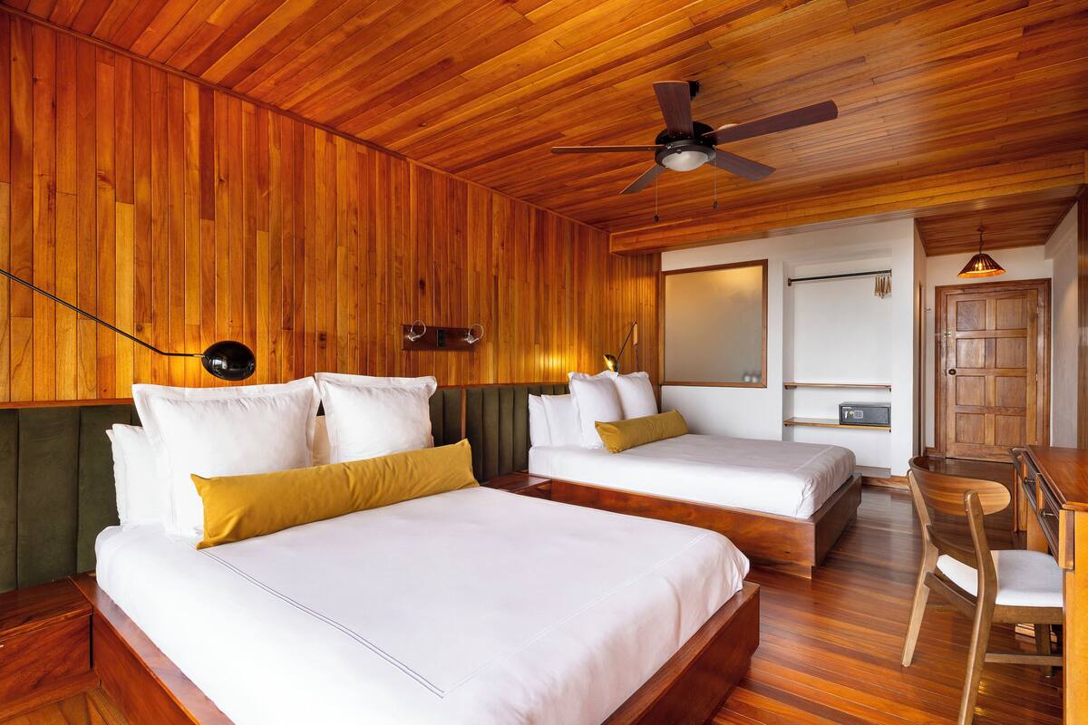 Une chambre d’hôtel avec deux lits, des murs en bois, un ventilateur de plafond, un bureau avec une chaise et un miroir.