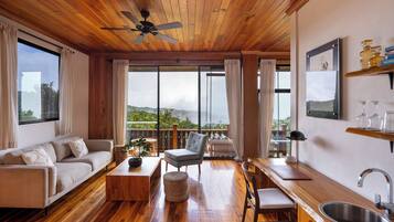 A spacious living area with a wooden ceiling, large windows, a sofa, a chair, a coffee table, and a kitchen area with a sink and shelves.