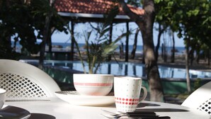 A table with two cups and a saucer, a fork, and a knife. The table is outdoors with a view of a building and trees. - Private Front Beach House (Huehuete)