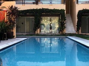 A swimming pool with a glass door entrance, surrounded by potted plants and a staircase leading to an upper level. - Apartamento com Piscina a Beira Mar (Cabo de Santo Agostinho)