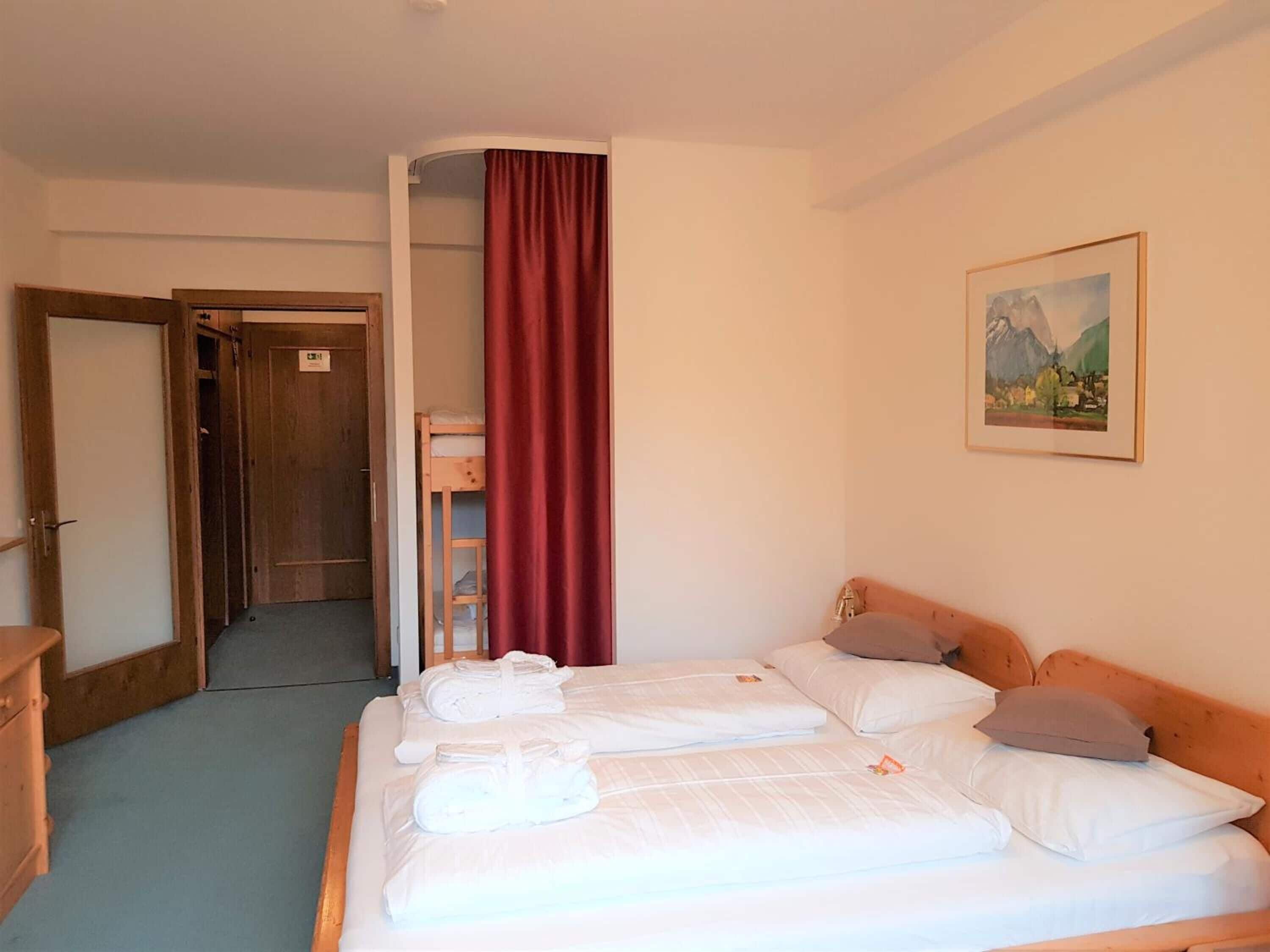 A hotel room with two beds, a wooden wardrobe, a door, a red curtain, and a painting on the wall.