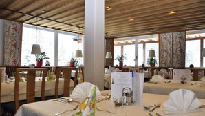 A restaurant with wooden ceiling beams, large windows with floral curtains, and tables set with white tablecloths and silverware. - Hotel Nockalm (Krems in Kaernten)