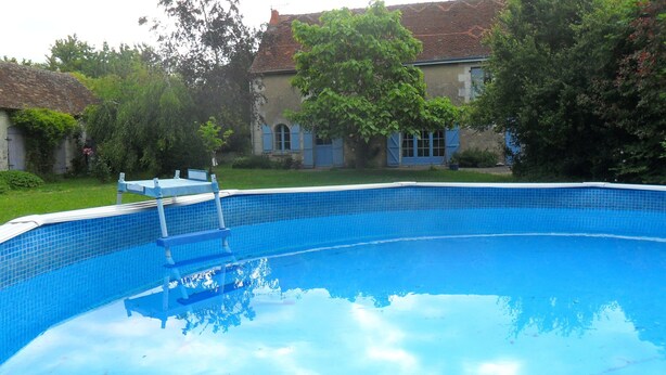 Ein Schwimmbecken mit Fliesenrand, umgeben von Grünpflanzen und einem Haus mit einem Ziegeldach.