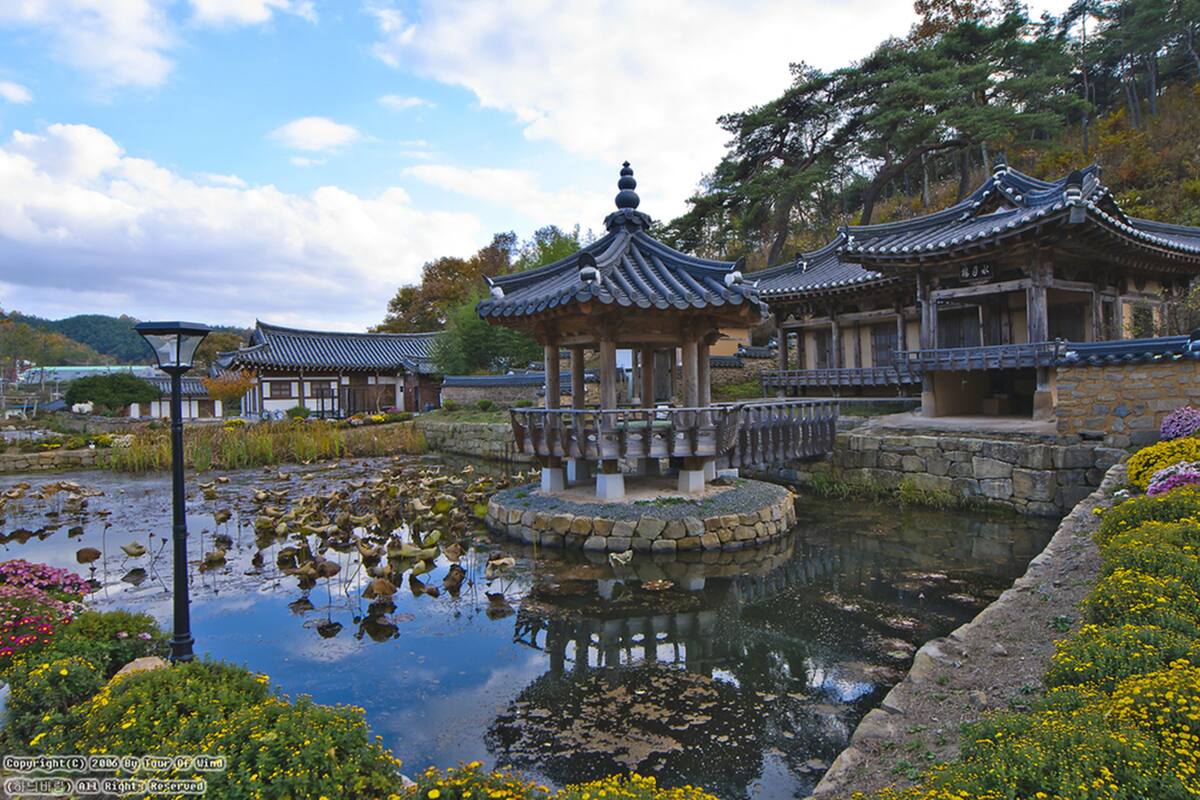 A traditional pavilion with a stone base and tiled roof, surrounded by a pond with lily pads and flowers.