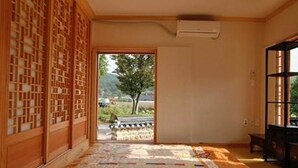 A room with a tatami mat floor, a sliding door leading to a view of a tree and road, and a traditional Japanese sliding door on the left. - Gwiae Traditional House (Yeongcheon)