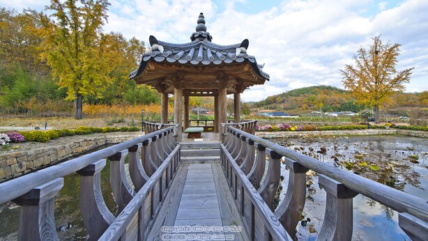 A wooden bridge leading to a traditional pavilion with a tiled roof, surrounded by autumn foliage and a pond. - Gwiae Traditional House (Yeongcheon)