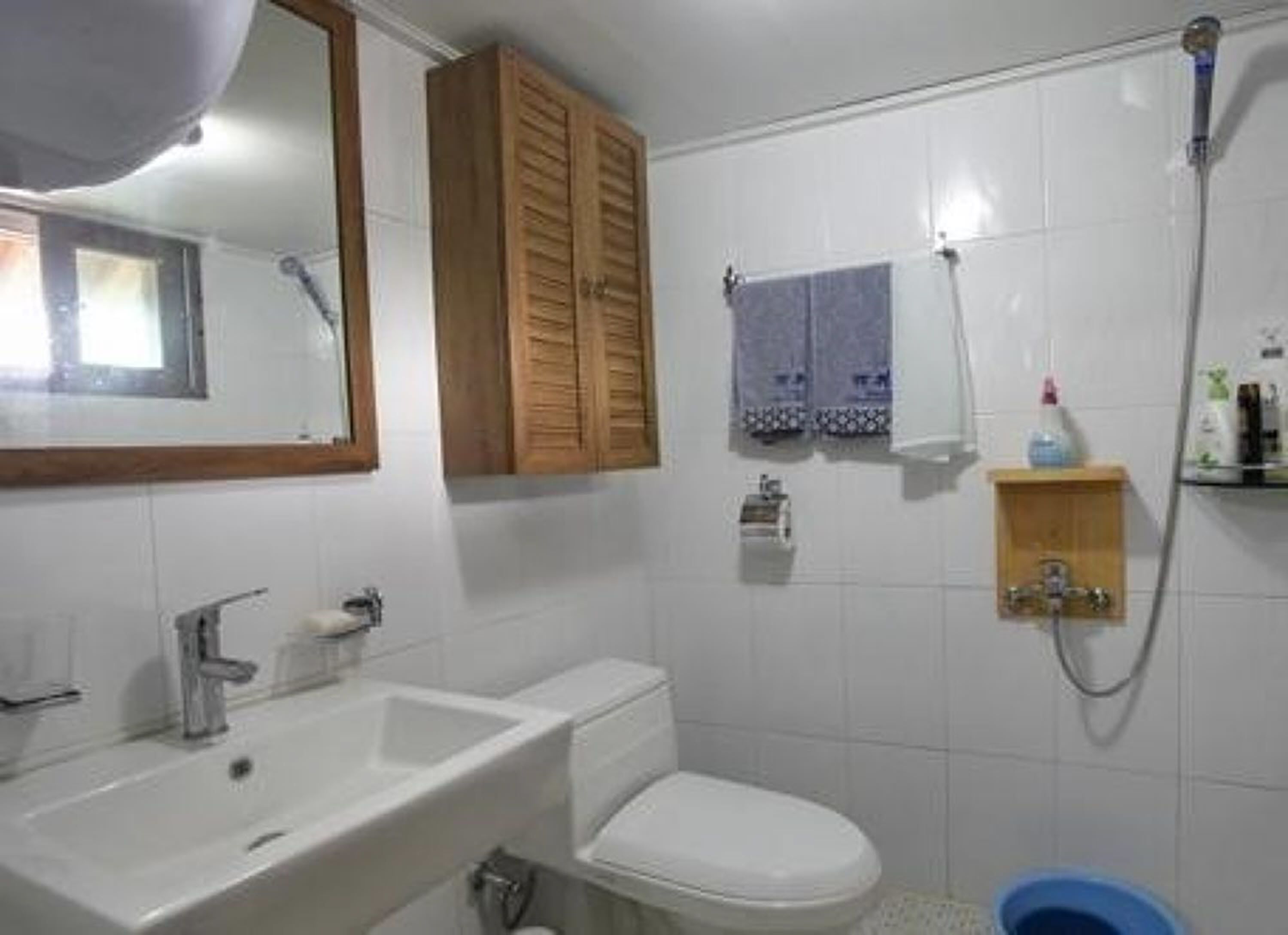 Une salle de bain avec un lavabo, des toilettes et une douche, le tout de couleur blanche. On trouve un meuble en bois au-dessus du lavabo et une serviette accrochée au mur.