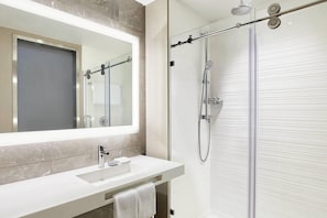 A modern bathroom with a glass shower enclosure, a white sink with a mirror above it, and a towel hanging on the wall. - AC Hotel Huntsville Downtown (Huntsville)