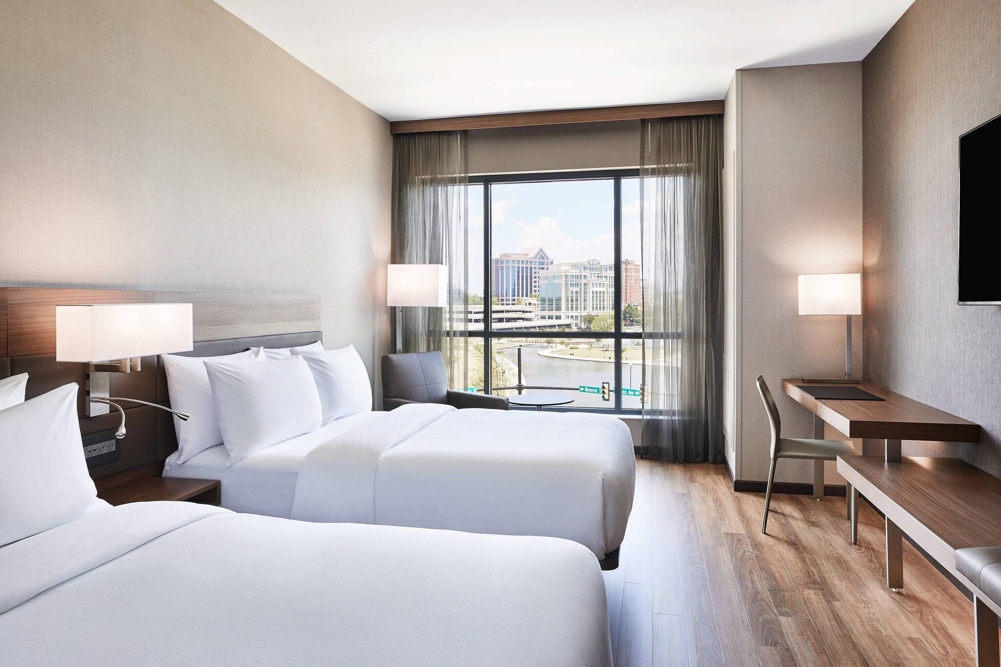 A hotel room with two beds, a desk, and a view of a cityscape.