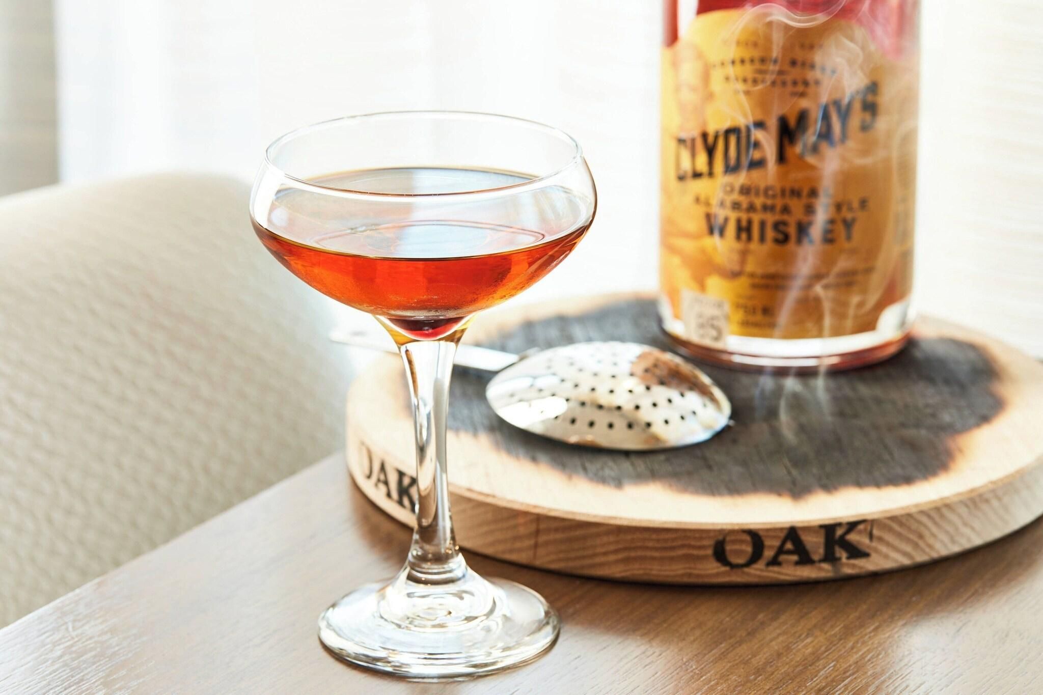 A whiskey glass with a drink on a wooden coaster, a bottle of whiskey, and a cookie on a wooden tray.