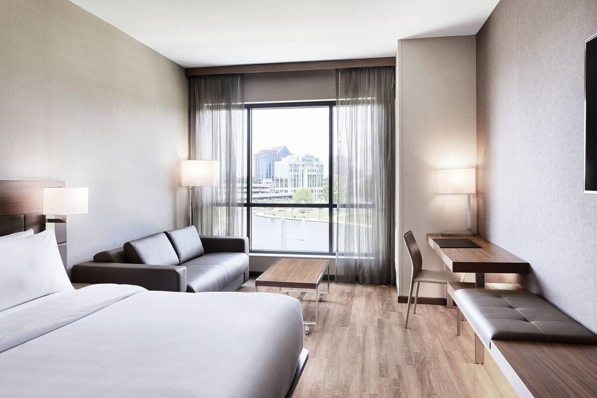 Une chambre d’hôtel moderne avec un grand lit, un canapé, un bureau et une chaise. Une grande fenêtre, dotée de rideaux, offre une vue sur la ville.