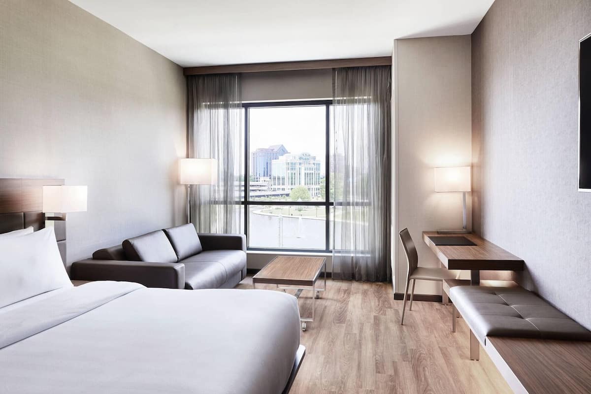 Moderni hotellihuone, jossa on suuri sänky, sohva, työpöytä ja tuoli. Huoneessa on suuri ikkuna, jossa on verhot, ja ikkunasta avautuu näkymä kaupunkiin.