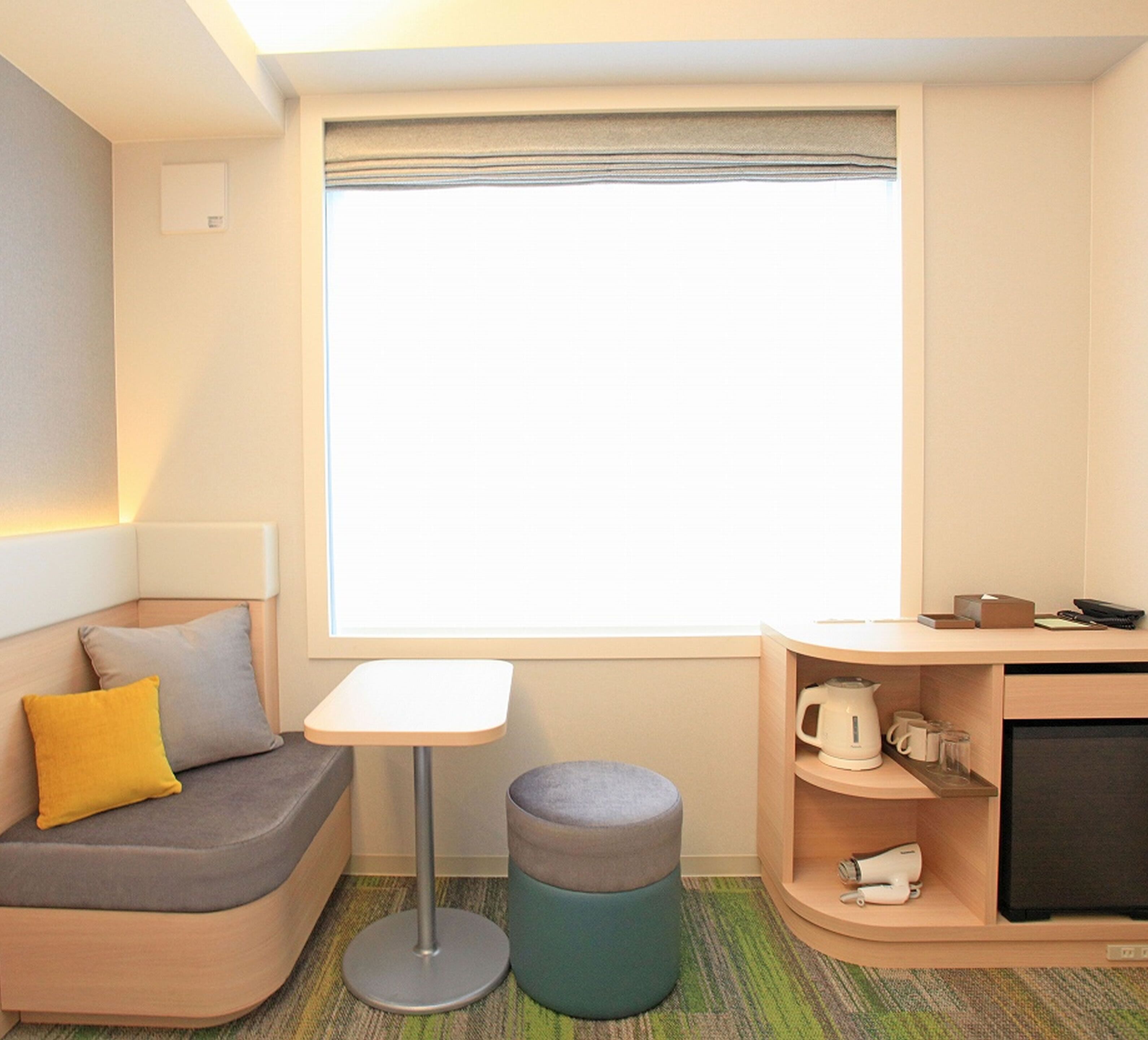 A compact hotel room with a sofa, a small table, a round ottoman, a kitchenette, and a large window.