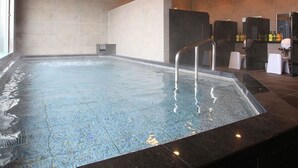 A large indoor swimming pool with a tiled floor, a metal pool ladder, and a wall with a built-in bench. - Urban Hotel Kyoto Gojo Premium (Kyoto)