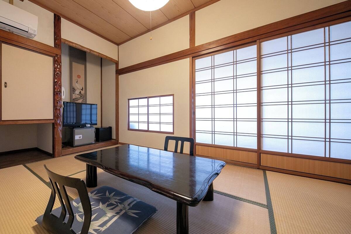 Une pièce de style japonais traditionnel, avec un sol recouvert de tatamis, une table basse, des chaises, un téléviseur et des portes coulissantes.
