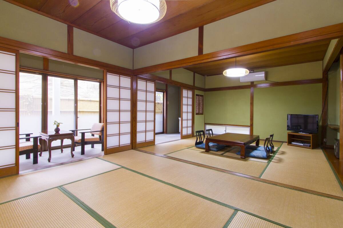 Une pièce de style japonais traditionnel, avec un sol recouvert de tatamis, des portes coulissantes, une petite table basse avec des coussins et un téléviseur.