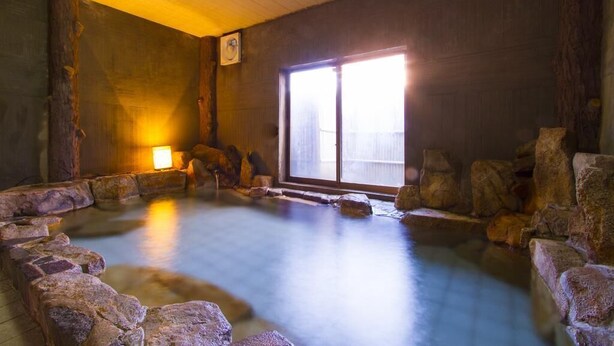 A traditional Japanese onsen with a wooden interior, a stone bath surrounded by rocks, and a sliding door leading to the outside. - Takezono Ryokan (Shimonoseki)