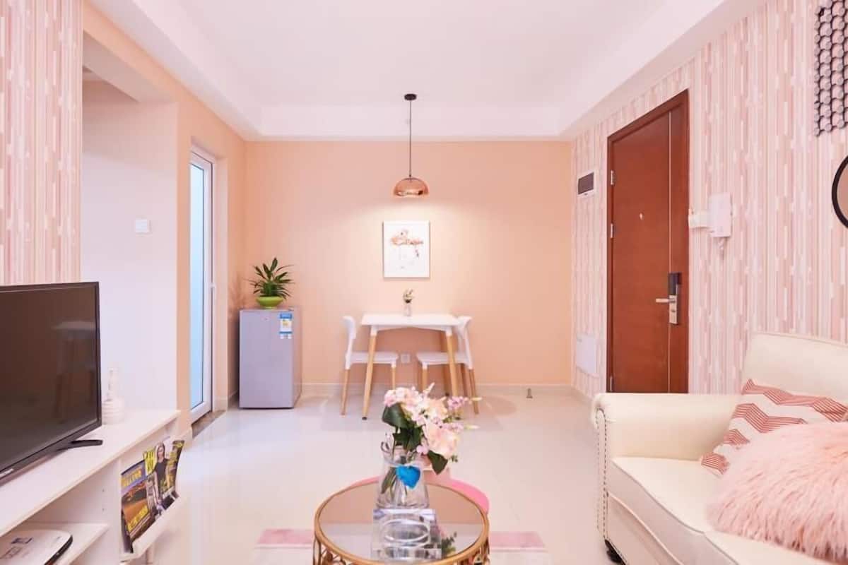 A modern living room with a flat-screen TV, a coffee table with a vase of flowers, a dining area with a white table and chairs, and a sofa with a fluffy pillow.