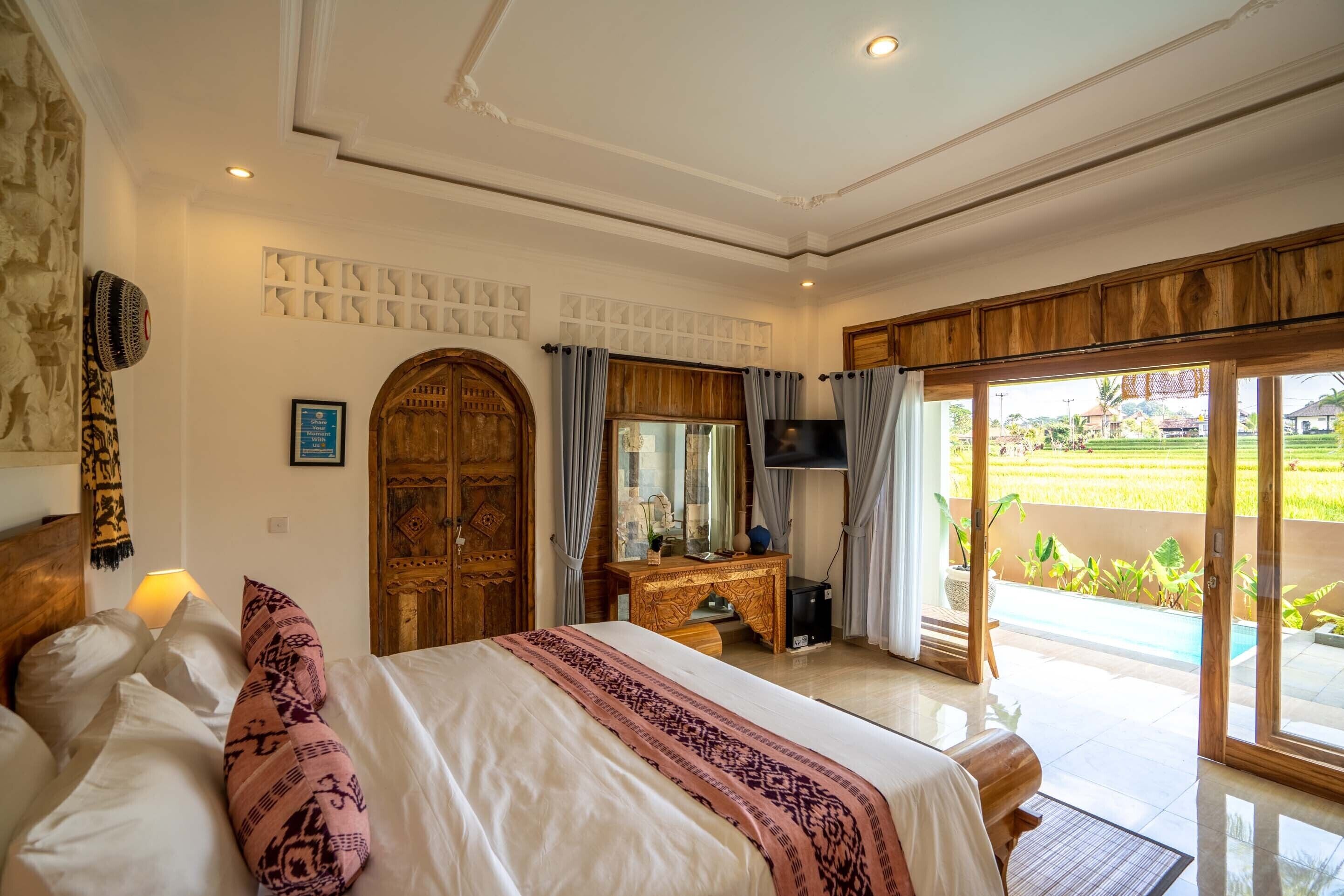 A bedroom with a large bed, wooden wardrobe, a desk with a TV, and a sliding glass door to a balcony.