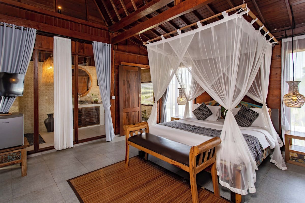 A wooden four-poster bed with white curtains, a television on a wooden stand, and a woven basket on a side table.
