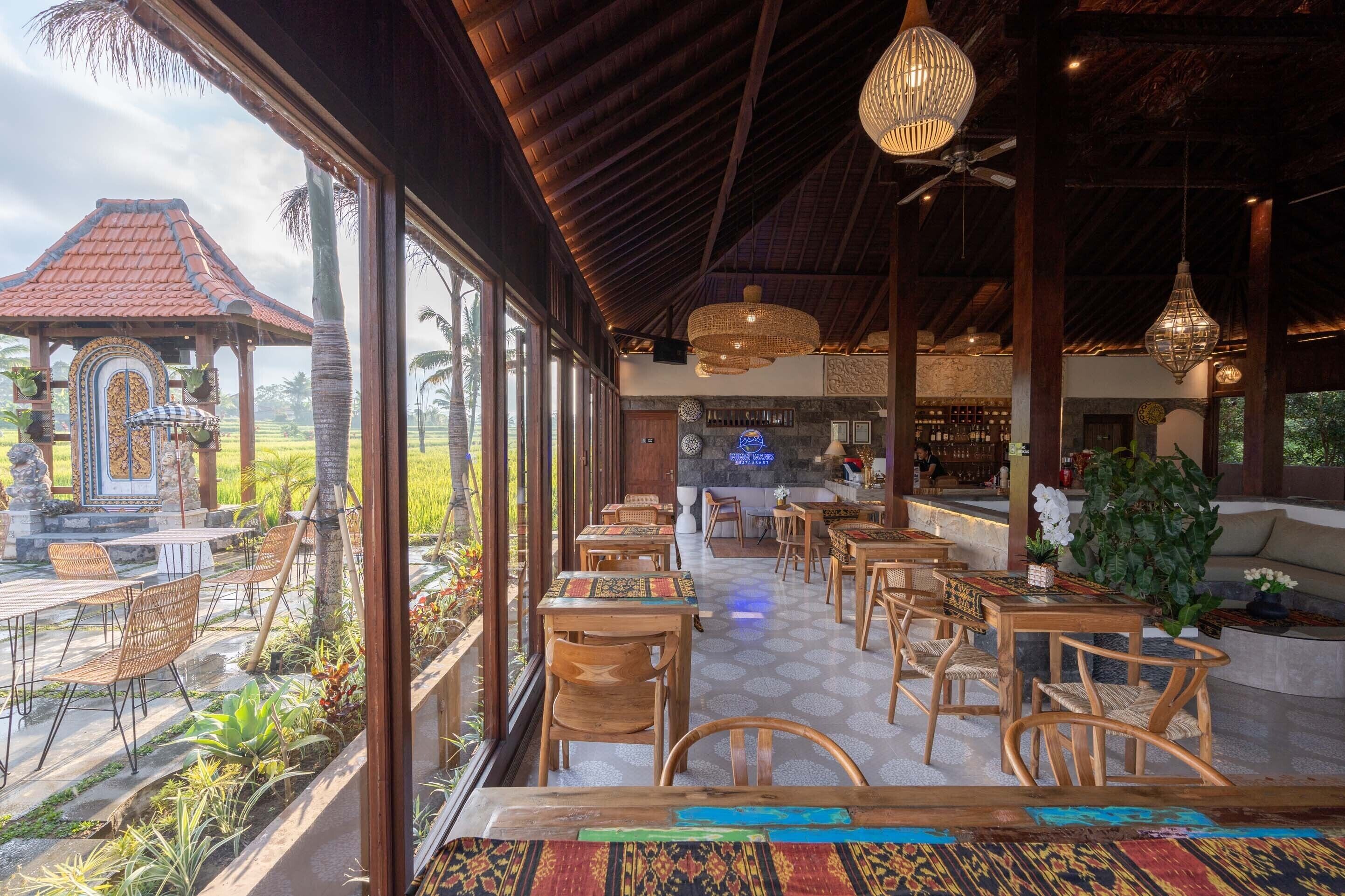A spacious indoor area with wooden tables and chairs, large windows offering views of a traditional building and palm trees, and a bar area with a stone counter.