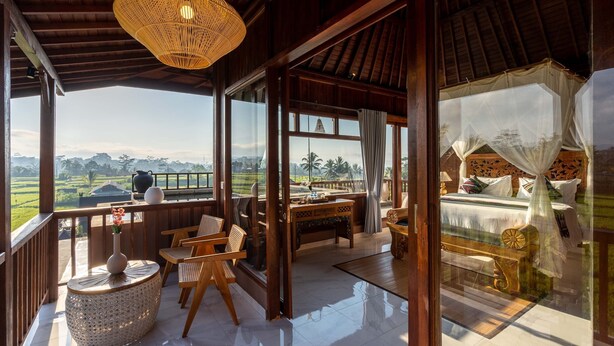 A room with a large window, a bed with a canopy, a wooden table, and a wicker chair.