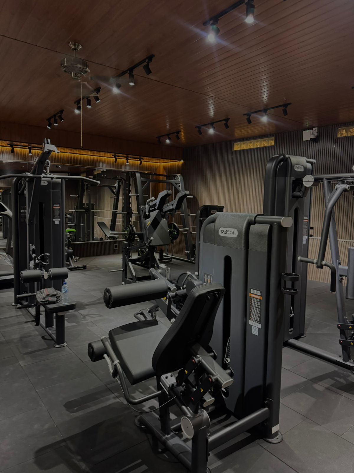 Un gimnasio con diversas máquinas de ejercicio, un ventilador de techo y una botella de agua sobre un banco.