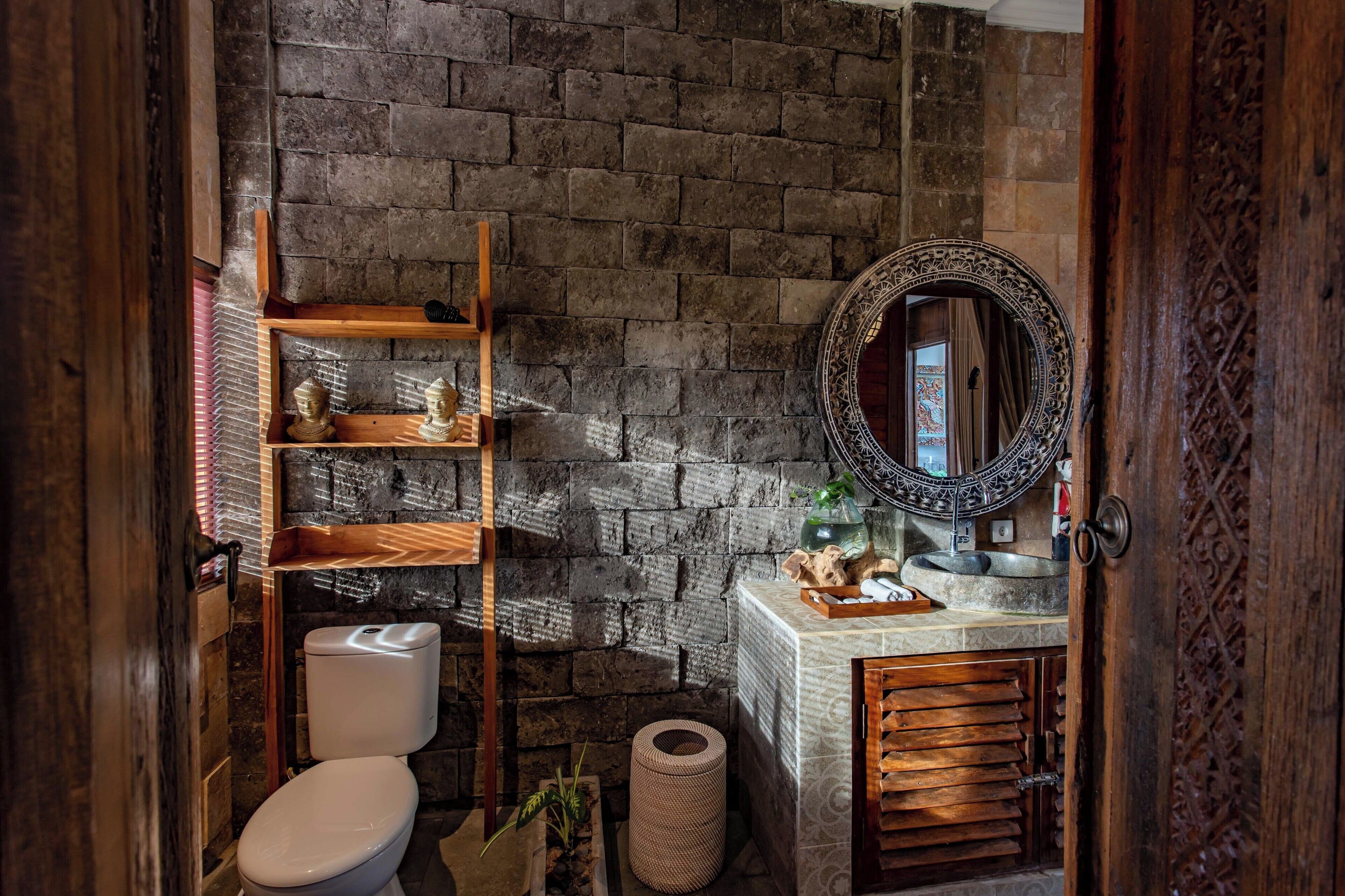 A bathroom with a brick wall, a wooden shelf with toiletries, a toilet, a sink with a decorative mirror, and a small plant.