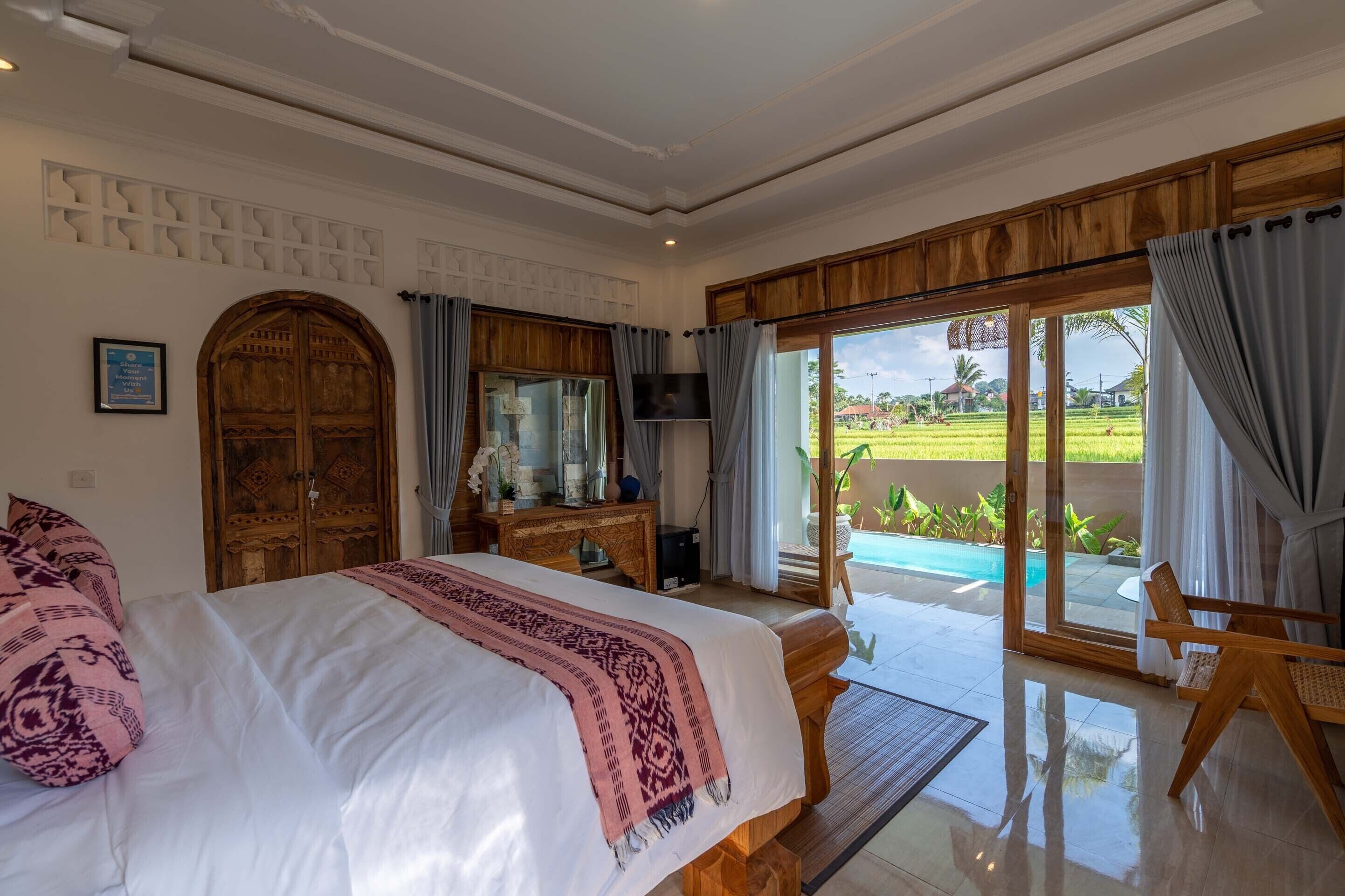 A bedroom with a large bed, wooden wardrobe, and a TV. It has a balcony with a view of a swimming pool and greenery.