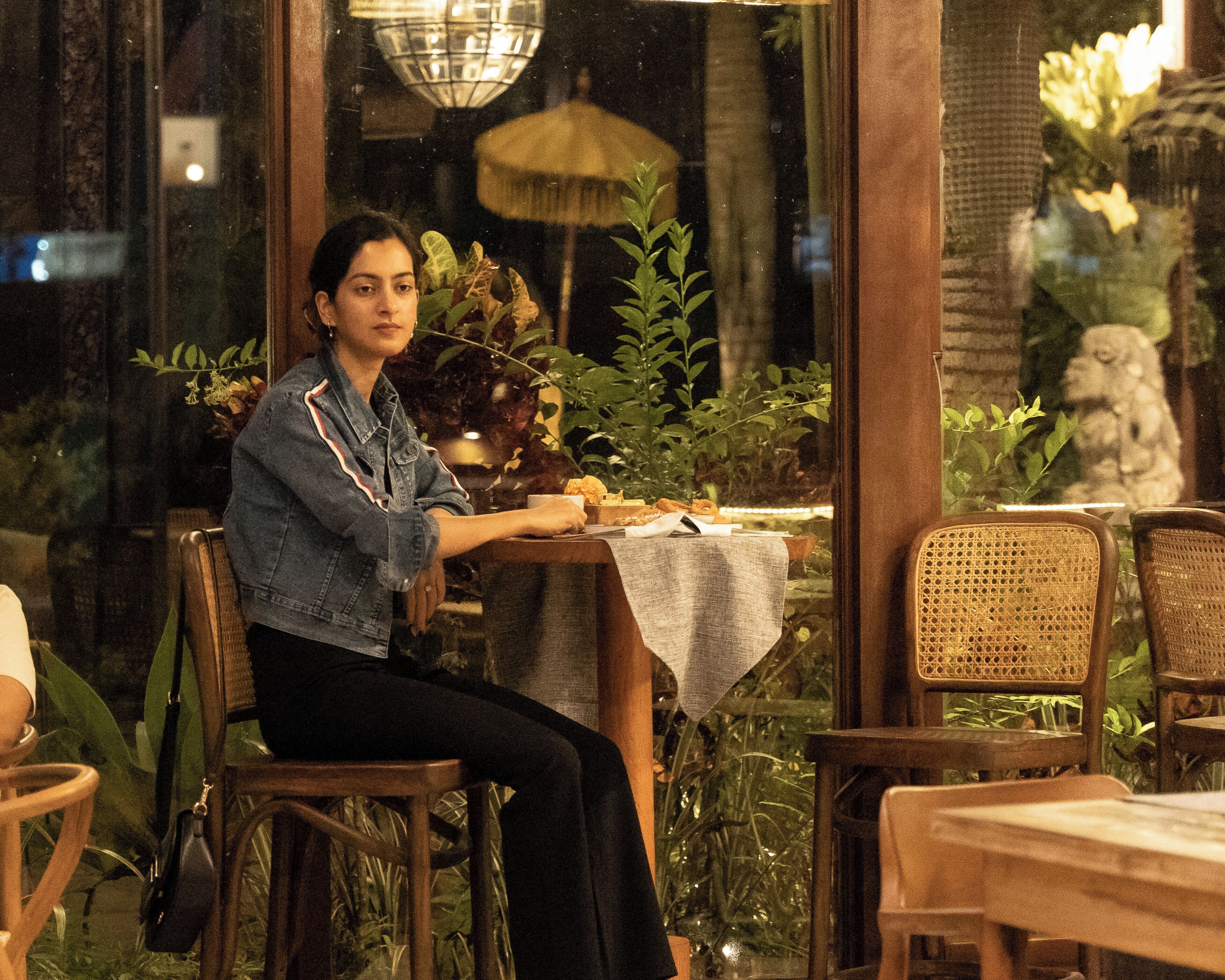 A person seated at a table with a plant, a lamp, and a statue in the background.