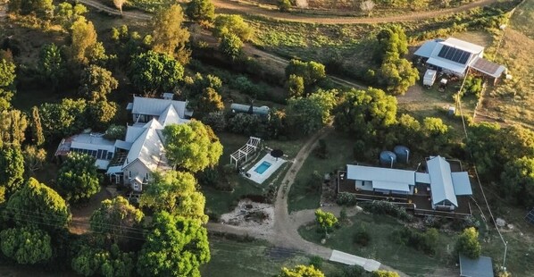 Vista aérea de una propiedad rural con varias edificaciones, incluyendo una casa principal con un garaje grande, una estructura más pequeña con paneles solares y una piscina.
