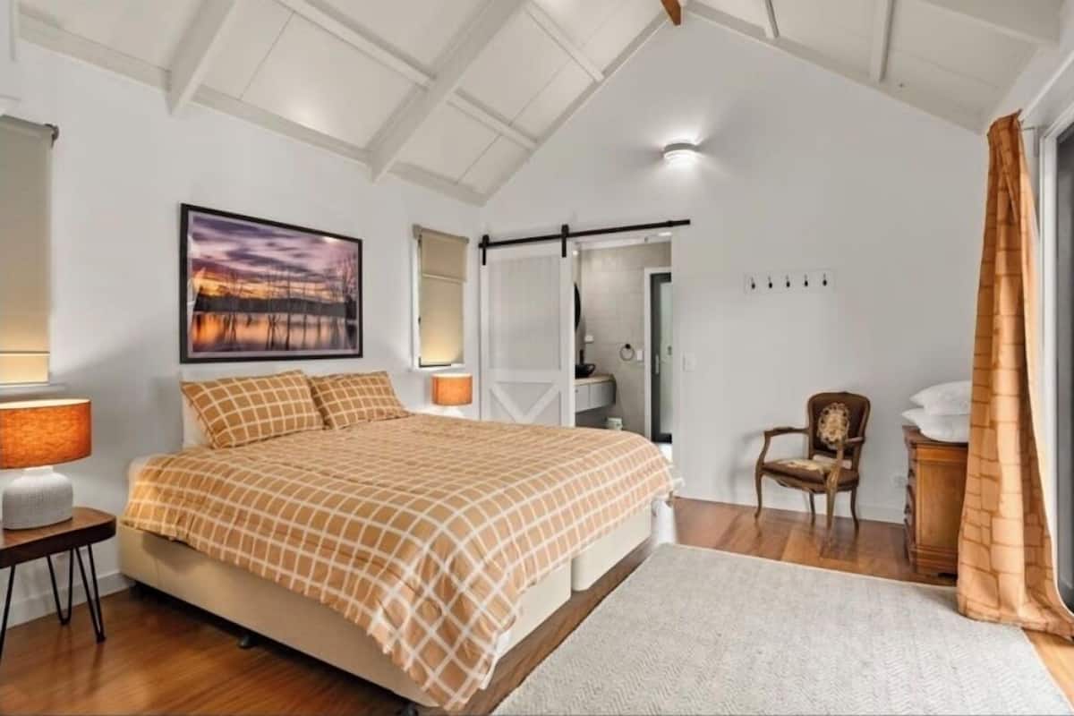 A bedroom with a bed, a nightstand, a chair, a window with curtains, and a painting on the wall.
