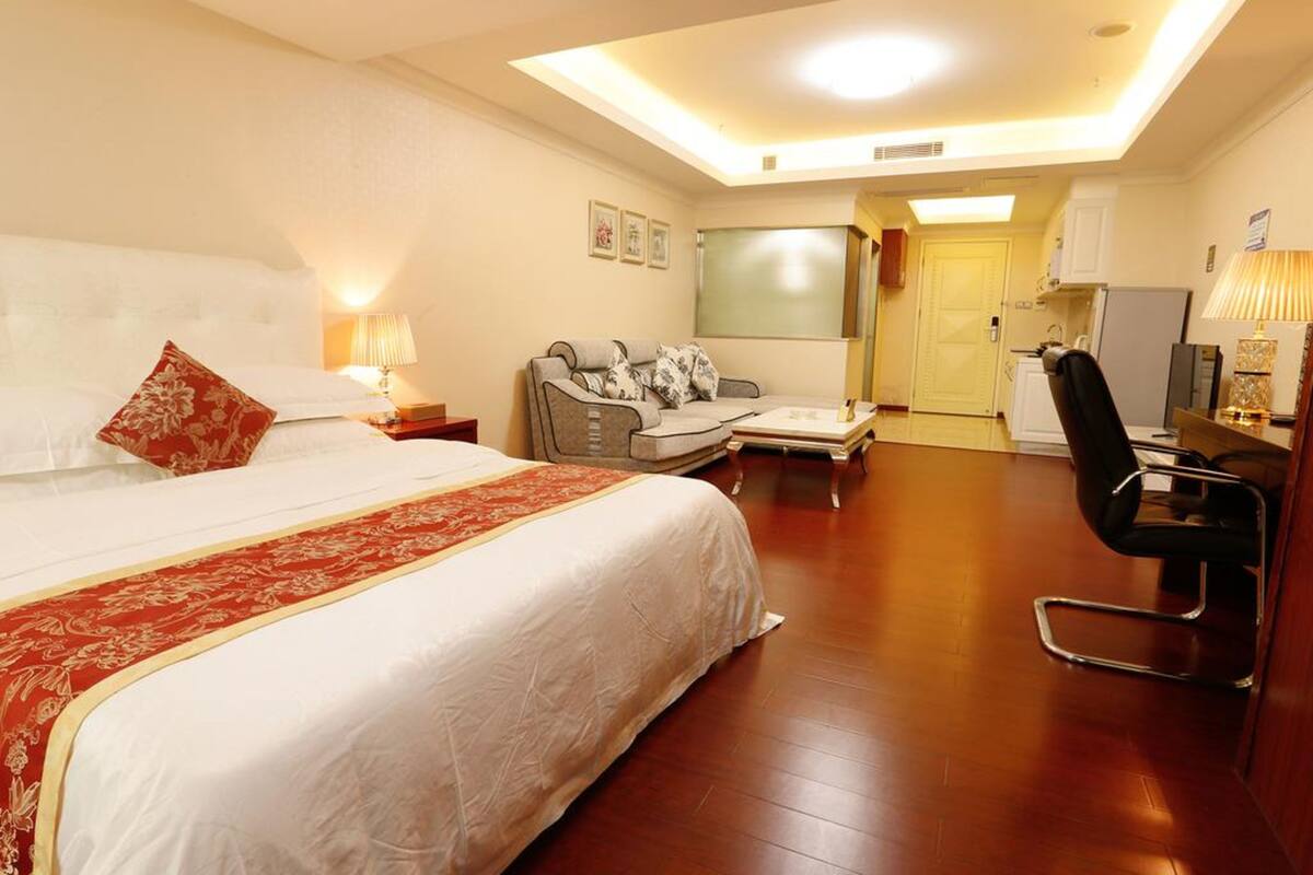 A hotel room with a large bed, a sofa, a desk, and a chair. There is a kitchen area with a refrigerator and a sink.