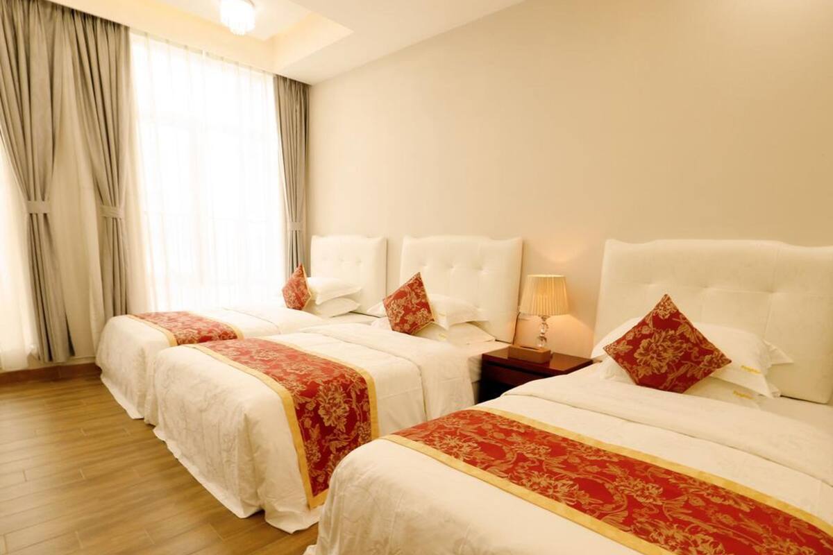 A hotel room with two beds, each with white bedding and red floral bedspreads. There is a nightstand with a lamp between the beds, and a large window with curtains.