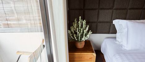 A bedroom with a bed, a wooden nightstand, a potted plant, and a window with blinds.
