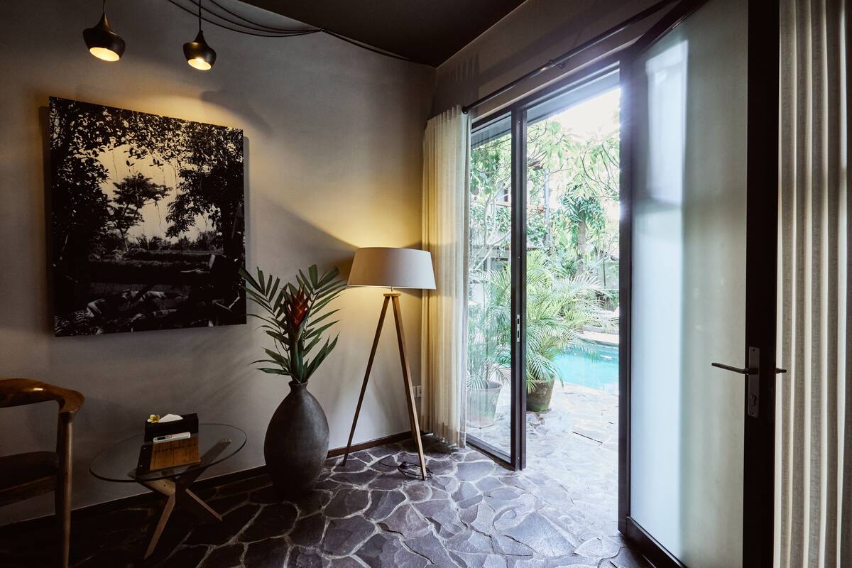 A room with a stone floor, a large window with curtains, a painting on the wall, a lamp, and a small table with a plant.