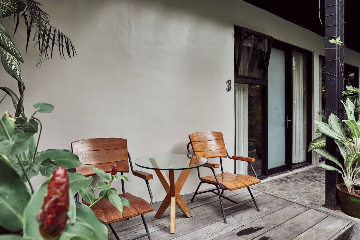 A small outdoor seating area with a glass-top table and wooden chairs, surrounded by potted plants and a building entrance.