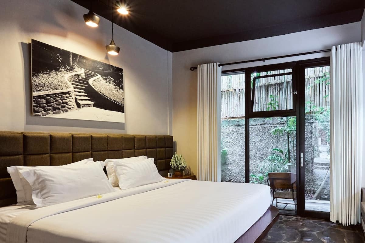A neatly made bed with white linens, a large framed picture above it, and a view of a garden through a glass door.