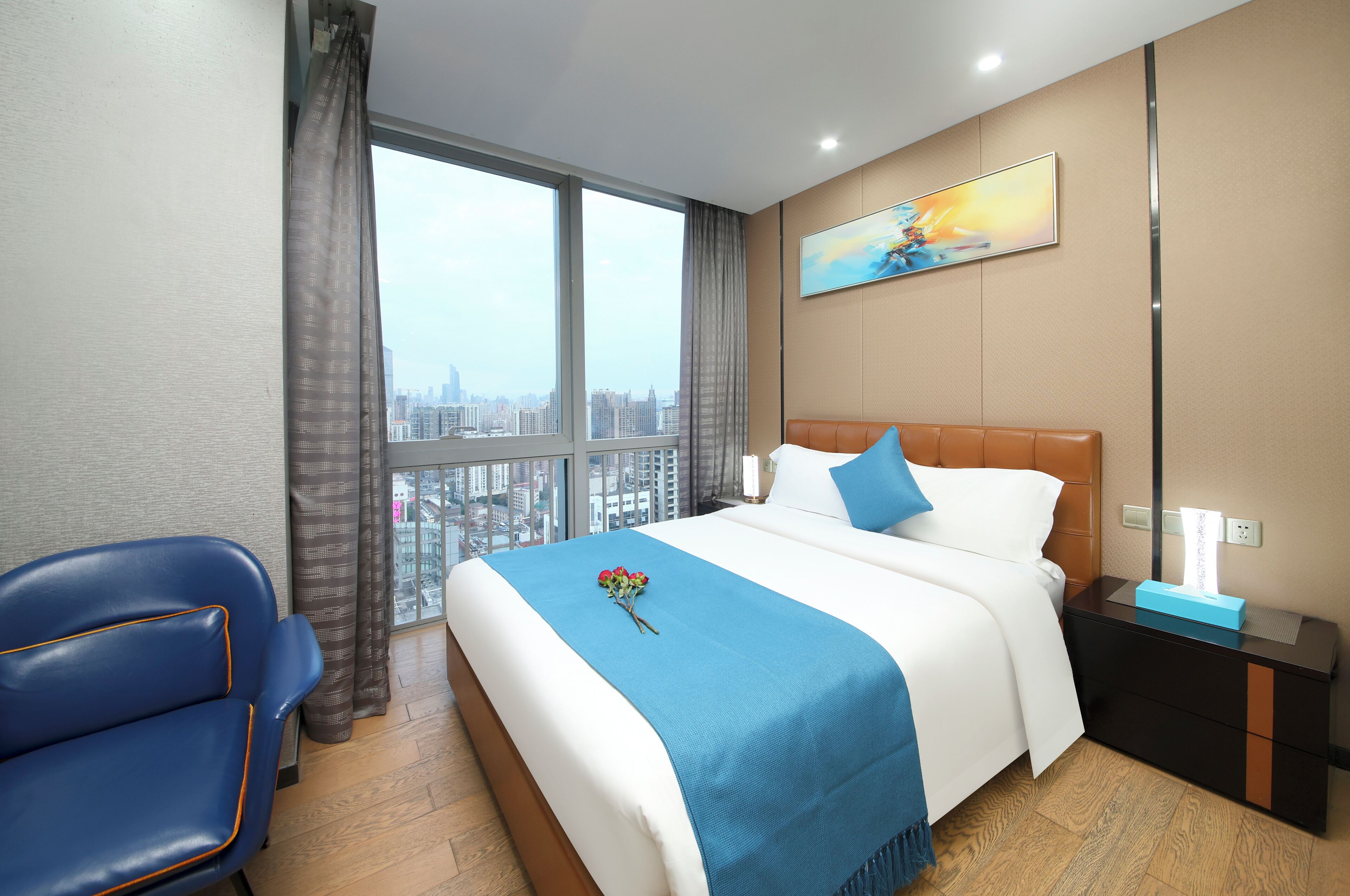 A hotel room with a large window, a bed with a blue bedspread, a blue chair, and a nightstand with a lamp.