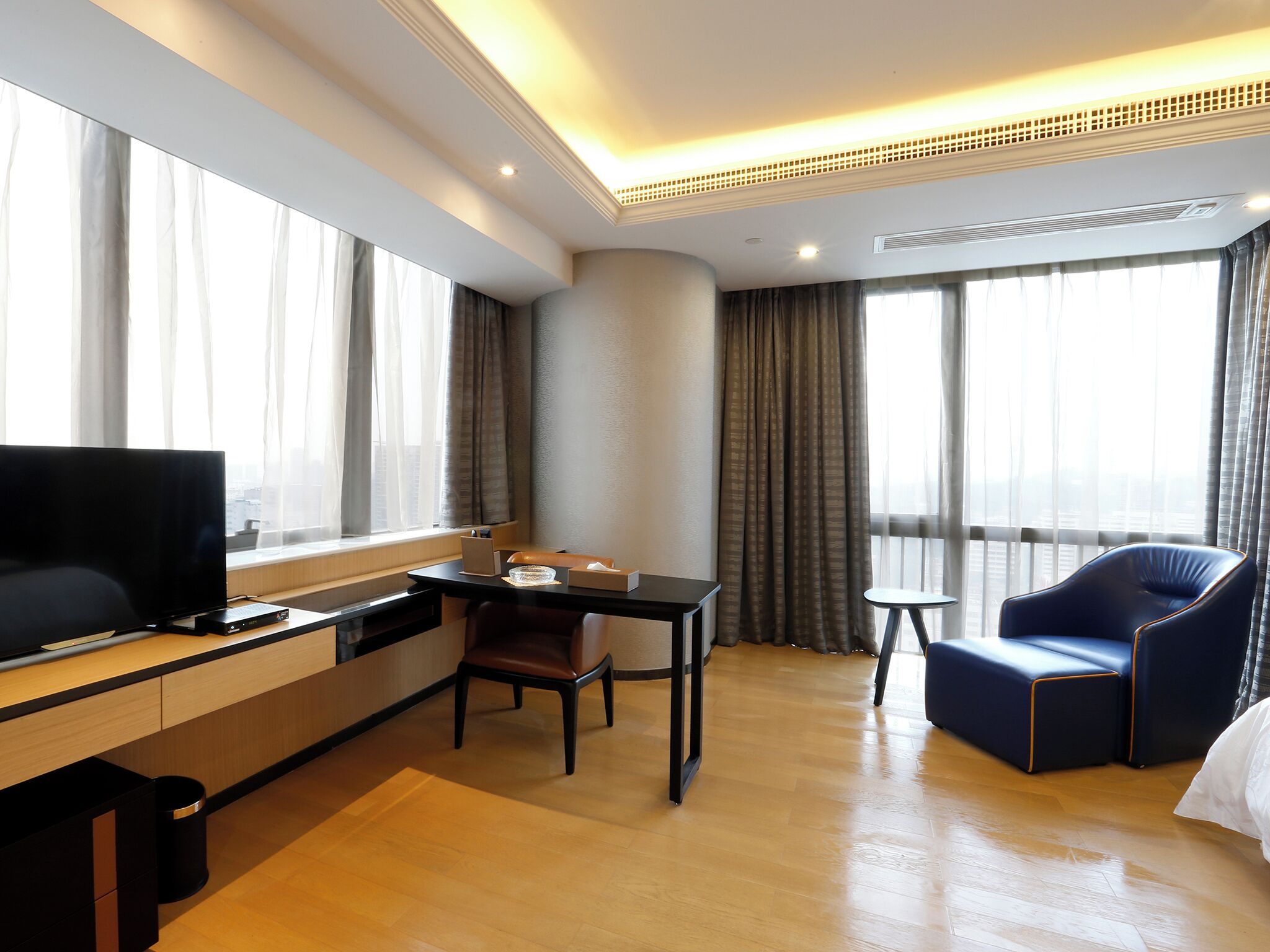 A hotel room with a flat-screen TV, a small dining table with chairs, a sofa, and a large window with curtains.