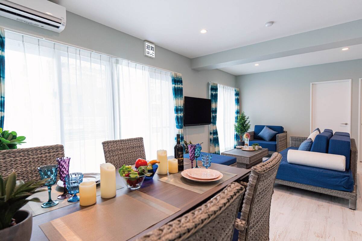 A modern living room with a dining area, featuring wicker furniture, a flat-screen TV, and a large window with sheer curtains.