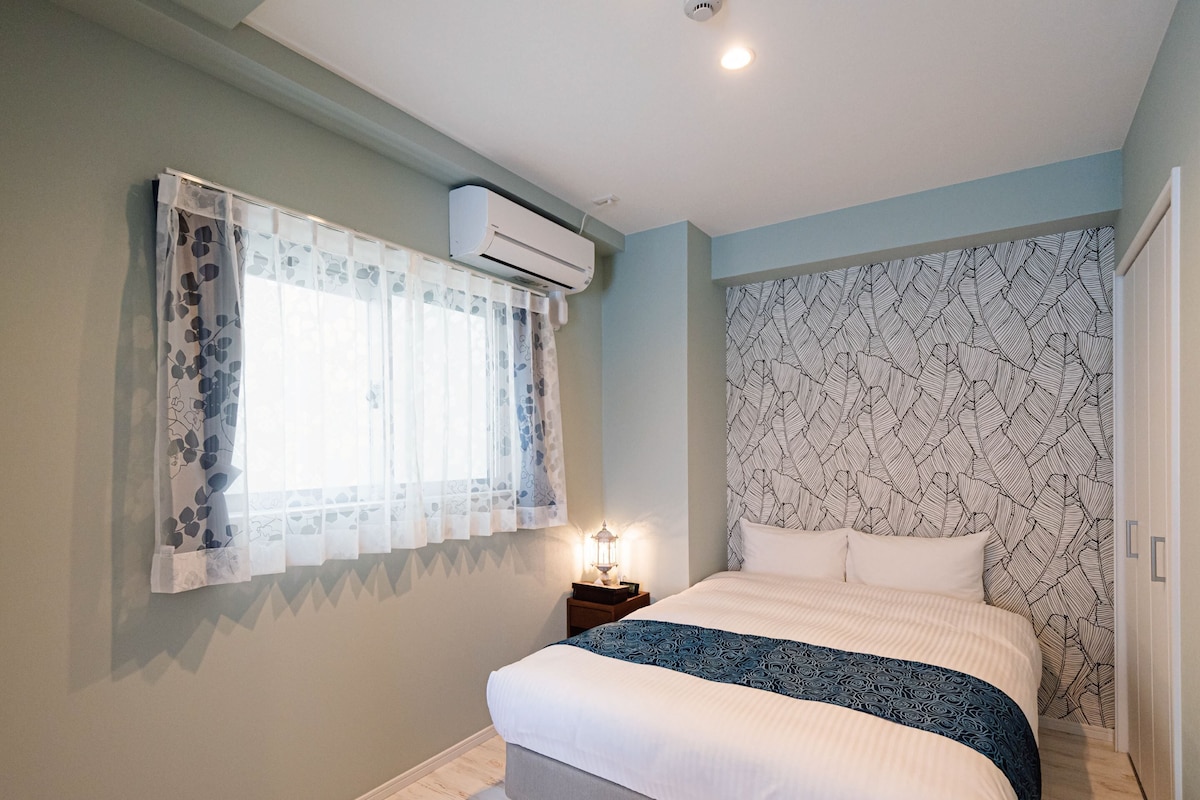 A bedroom with a bed, a window with sheer curtains, an air conditioner, a patterned wallpaper, and a nightstand with a lamp.