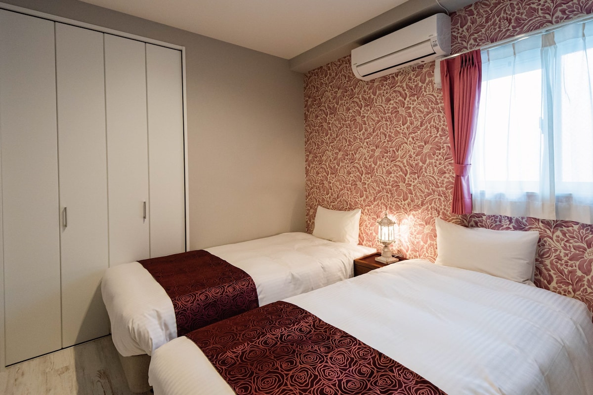 A hotel room with two beds, a wardrobe, an air conditioning unit, and a window with curtains.