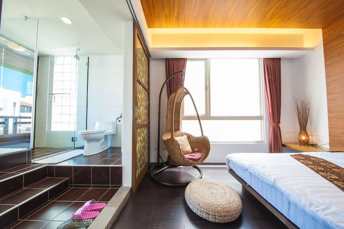 A modern hotel room with a large window, a hanging chair, a bed, and a bathroom with a shower and toilet.