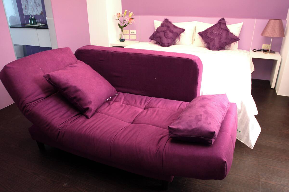 A purple sofa with matching cushions in a room with a bed and a nightstand.