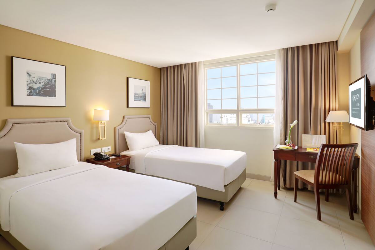 Ein Hotelzimmer mit zwei Betten, einem Schreibtisch mit Stuhl, einem Fenster mit Vorhängen und gerahmten Bildern an der Wand.