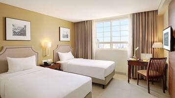 A hotel room with two beds, a desk with a chair, a window with curtains, and framed pictures on the wall.