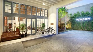 A building entrance with a large glass door, potted plants, a bicycle, and a neon sign reading "KOKOON". - Kokoon Hotel Surabaya (Surabaya)