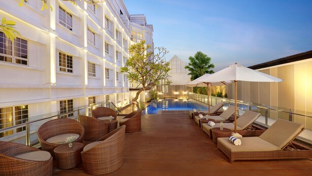 A rooftop pool area with lounge chairs and wicker furniture, surrounded by a white building with large windows and a clear sky in the background. - Kokoon Hotel Surabaya (Surabaya)