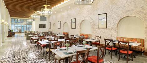Un restaurante espacioso con techos altos, paredes de ladrillo y un suelo con diseño. Las mesas están cubiertas con manteles blancos y sillas rojas.